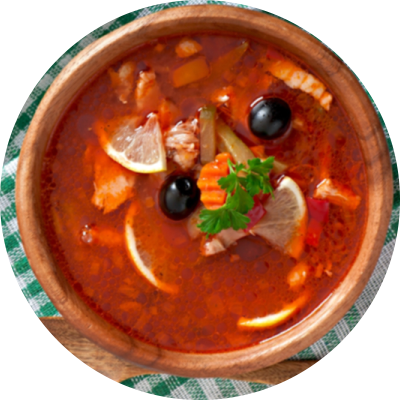 Eine Holzschüssel mit leuchtend roter Suppe mit Zitronenscheiben, schwarzen Oliven, Karotten und frischer Petersilie auf einem grün-weiß karierten Tuch.