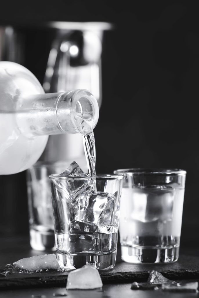 Eine Glasflasche gießt klare Flüssigkeit in ein mit Eiswürfeln gefülltes Glas, umgeben von zwei weiteren ähnlichen Gläsern. Die Szene spielt vor einem dunklen Hintergrund, im Hintergrund ist ein verschwommener metallischer Gegenstand zu sehen.