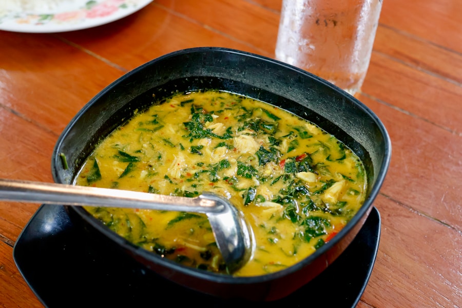 Eine schwarze Schale, gefüllt mit gelber Currysuppe mit Blattgemüse und Gemüse, in der ein Metalllöffel ruht. Die Schüssel steht auf einem passenden schwarzen Teller auf einem Holztisch, daneben ein Glas Wasser.