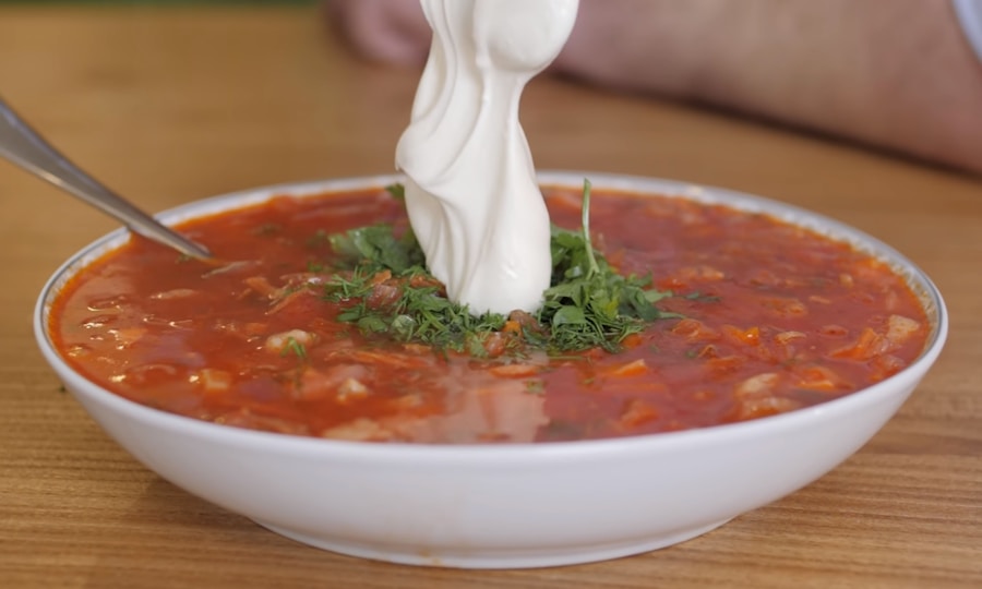 Eine Schüssel mit roter Suppe, wahrscheinlich Borschtsch, garniert mit frischen gehackten Kräutern und einem großen Klecks saurer Sahne. Ein Löffel ruht in der Schüssel, die auf einem Holztisch steht.