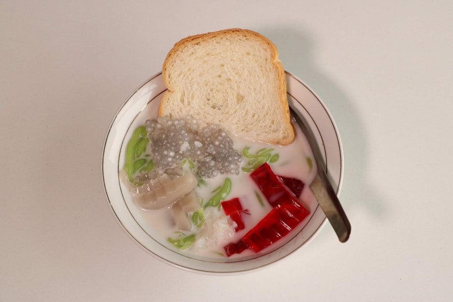 Eine Schale mit dem farbenfrohen indonesischen Dessert Cendol mit grünem Gelee, rotem Gelee, Palmenkernen, Tapiokaperlen, Kokosmilch und einer Scheibe Weißbrot obenauf, mit einem Löffel in der Schale.